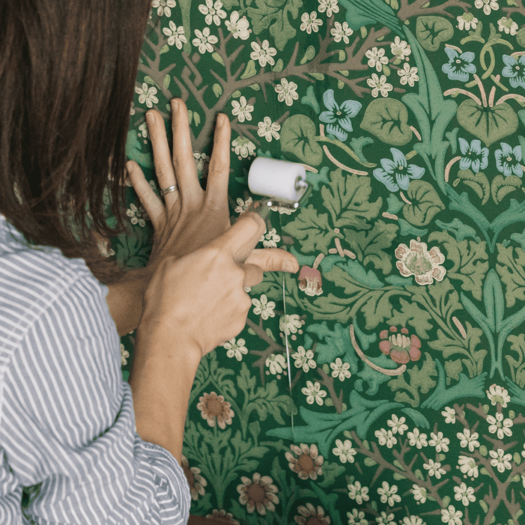 グリーンを基調にしたボタニカル柄の壁紙を、小さなローラーで圧着している様子。葉や花の繊細な模様が描かれたクラシックなデザインで、手で押さえながら丁寧に貼り付けているシーン。