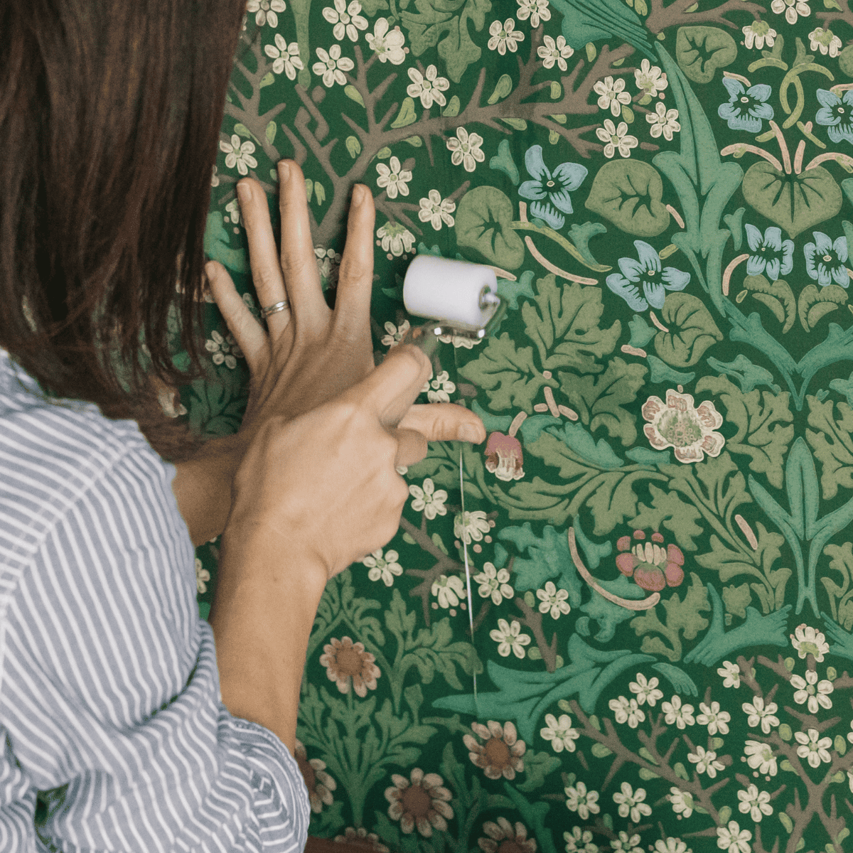 グリーンを基調にしたボタニカル柄の壁紙を、小さなローラーで圧着している様子。葉や花の繊細な模様が描かれたクラシックなデザインで、手で押さえながら丁寧に貼り付けているシーン。