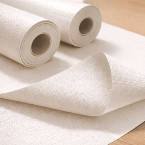 白系のビニールクロス（塩ビ）を木目の作業台に広げ、ロール状の壁紙とめくった断面から、塩ビ素材特有の均一な質感と施工しやすさが伝わるイメージ写真