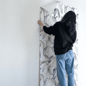 壁に貼られた既存の壁紙の上から、花柄の新しい壁紙を重ねて貼ろうとしている女性の後ろ姿。重ね貼りDIYの作業イメージ。
