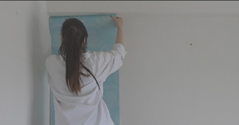 脚立に乗り、壁の上から青い柄の壁紙を貼り始めている様子。位置を合わせながら慎重に貼り付けている。