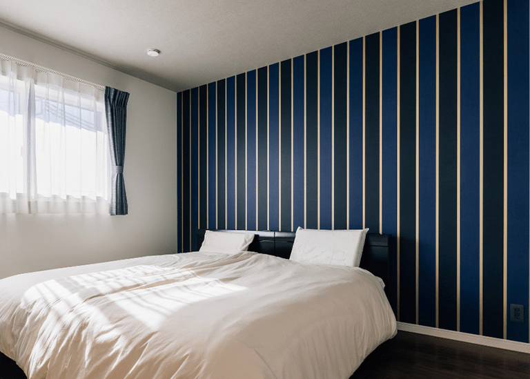 ネイビーの縦ストライプ壁紙をアクセントにした寝室。白い寝具と窓からの光が、落ち着いた雰囲気を作り出しているインテリア例。