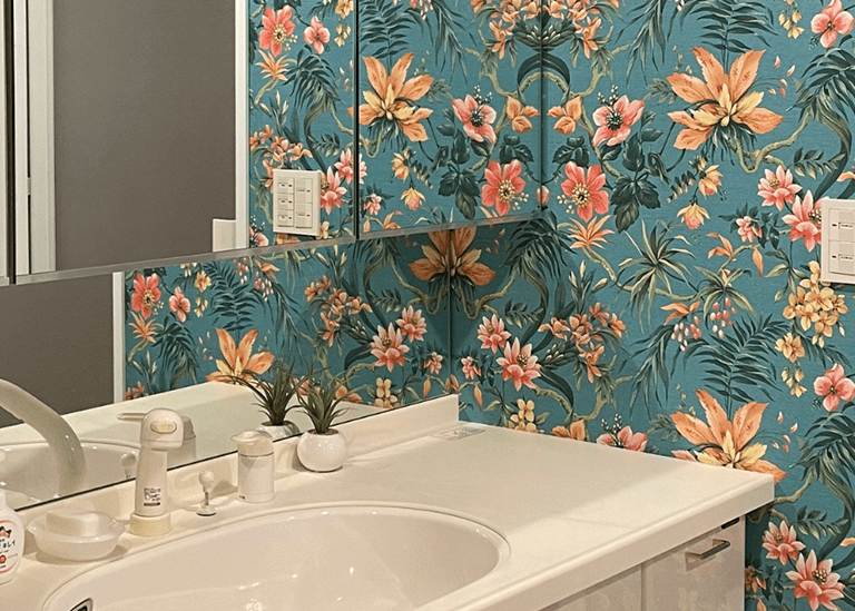 洗面所のアクセントクロス事例。ブルー系の壁紙を鏡まわりに使い、オレンジやピンクの花柄が映えるデザインと、白ベースの壁紙を並べて比較しているインテリア写真。
