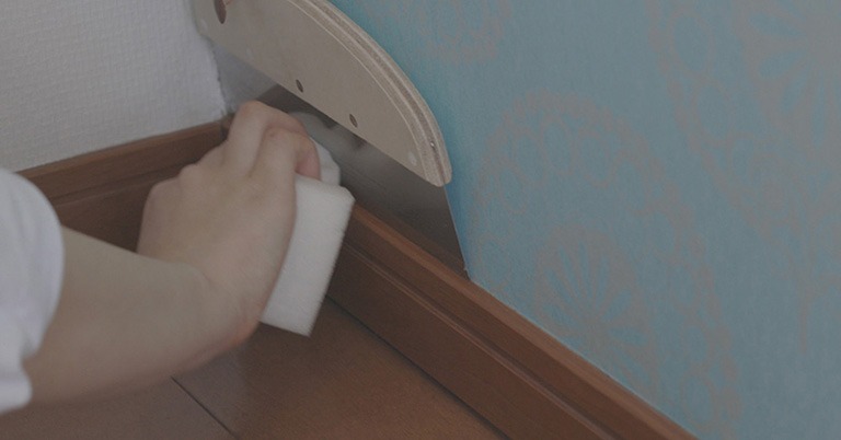 壁紙を貼ったあと、巾木についたのりをスポンジで丁寧に拭き取っている様子。壁紙が濡れないように地ベラで保護している。