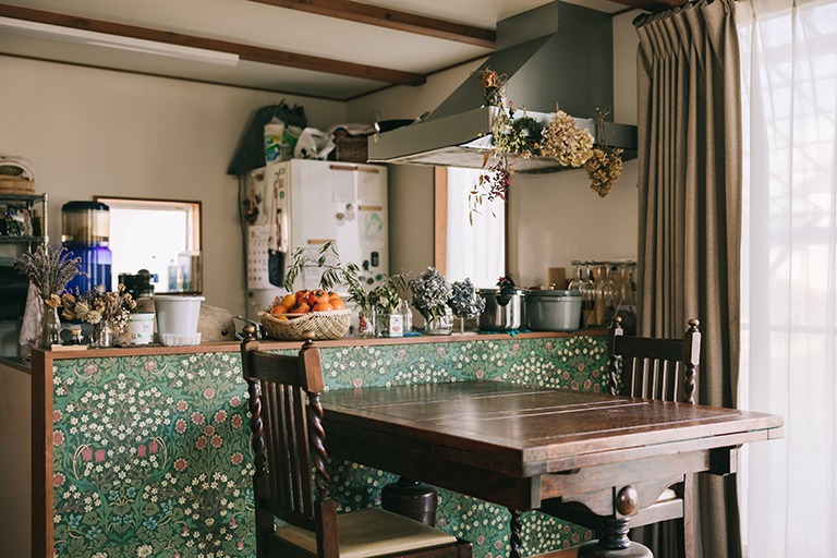 キッチンとダイニングをつなぐ腰壁にウィリアム・モリスのブラックソーン柄壁紙を用いたインテリア。深いグリーンの草花模様が木製テーブルや椅子、ドライフラワーの装飾と調和し、落ち着きと温かみのあるクラシカルな空間を演出している。