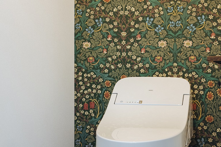 トイレ背面の壁にウィリアム・モリスのブラックソーン柄壁紙をアクセントとして使用した空間。深いグリーンを基調に小花や草木が描かれ、白いタンクレストイレとのコントラストが映える、落ち着きのあるクラシカルなインテリア。