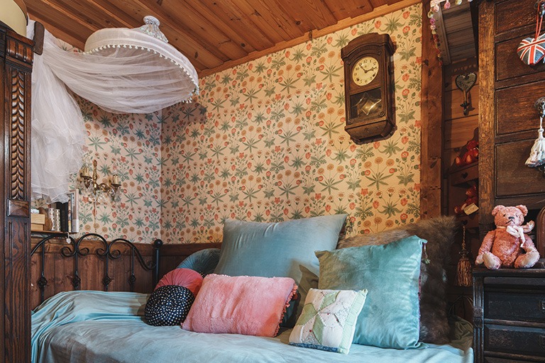 ウィリアム・モリスのデイジー柄壁紙を用いたベッドルームインテリア。木張りの天井とアンティーク調の家具に囲まれ、淡いブルーやピンクのクッションが重ねられた、温かみと物語性を感じるクラシカルな空間。
