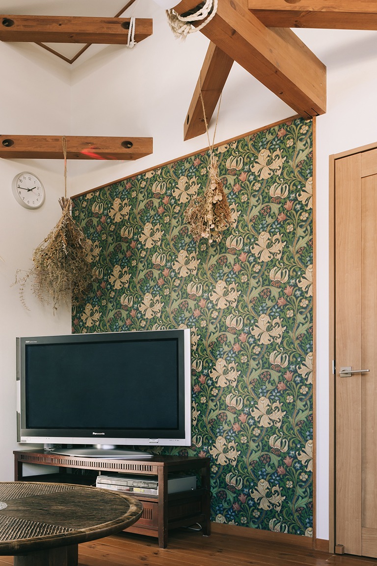 リビングのアクセントウォールにウィリアム・モリスのゴールデンリリー柄壁紙を使用したインテリア。木の梁が現しになった天井と、テレビボードやドライフラワーが調和し、深みのあるグリーンと草花模様が映えるクラシカルで温かみのある空間。