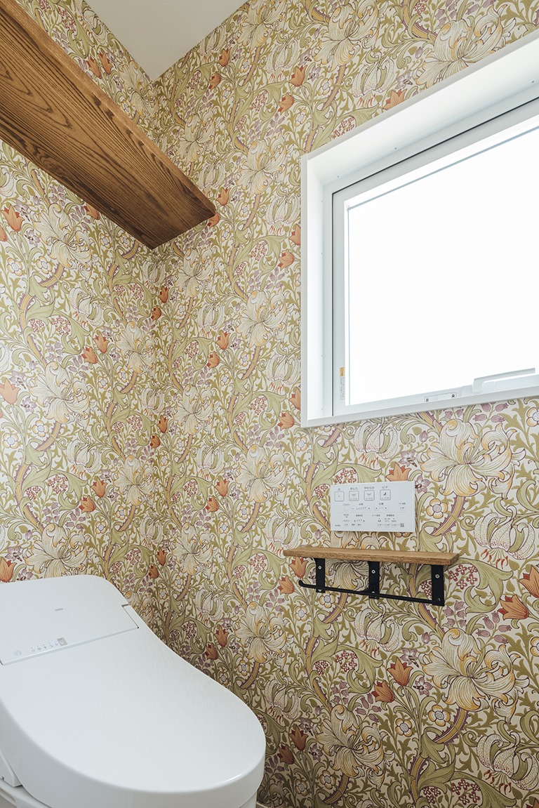 ウィリアム・モリスのゴールデンリリー柄壁紙を全面に使用したトイレ空間。アイボリーとイエローを基調とした草花模様に、白いタンクレストイレと木製棚、小窓が組み合わさり、明るく上品でクラシカルな雰囲気を演出している。