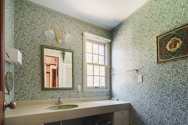洗面室の壁全面にウィリアム・モリスのシスル柄壁紙を使用したインテリア。ブルーグリーンを基調とした繊細な植物模様に、クラシカルなミラーと照明、窓からの自然光が調和し、上品で落ち着きのある空間を演出している。