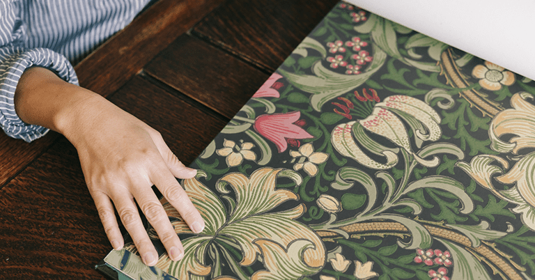 木製のテーブルの上で、人物の左手がウィリアム・モリス風の植物文様が描かれた壁紙サンプルに触れており、緑を基調に花や葉が緻密に描かれた装飾的なデザインが広がっている様子