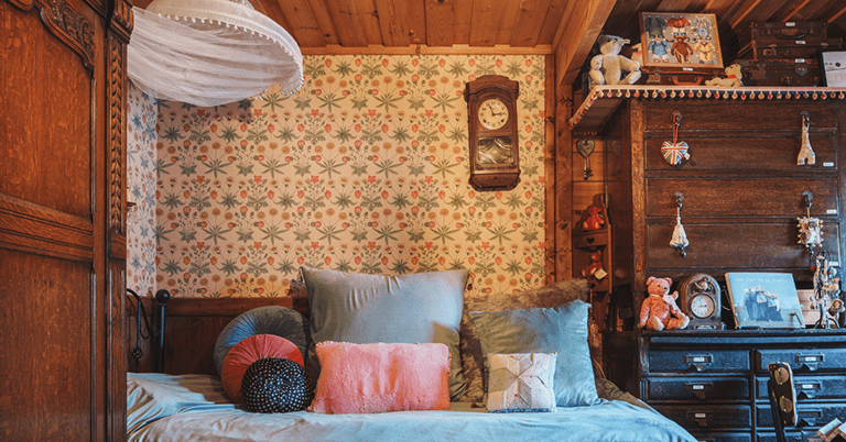 木の温もりを感じる寝室のインテリアで、ウィリアム・モリス風の小花と葉の壁紙がベッド背面に貼られ、クッションを並べたベッド、アンティーク調の家具や時計、ぬいぐるみや雑貨が並ぶ落ち着いた居心地のよい空間の様子