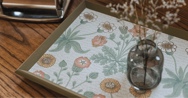 木製テーブルの上に置かれたトレーの底面に、ウィリアム・モリス風の草花文様が描かれており、淡いグリーンとピンク、ベージュの花柄が広がる中、手前には小さなガラス瓶に挿したドライフラワーが置かれた、ナチュラルで落ち着いた暮らしのワンシーン