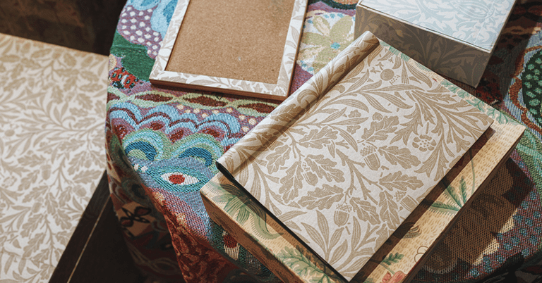 ウィリアム・モリス調の植物柄壁紙サンプルやノートが、色鮮やかな織物のテーブルクロスの上に重ねて置かれているコーディネートイメージ。ベージュ系のボタニカルパターンを中心に、落ち着きと装飾性を感じさせるインテリア素材。