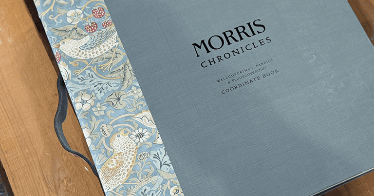 木製テーブルの上に置かれた『MORRIS CHRONICLES』と題されたコーディネートブックの表紙。落ち着いたブルーグレーの布張り表紙に、左側にはウィリアム・モリス調の鳥と草花の装飾柄があしらわれている。