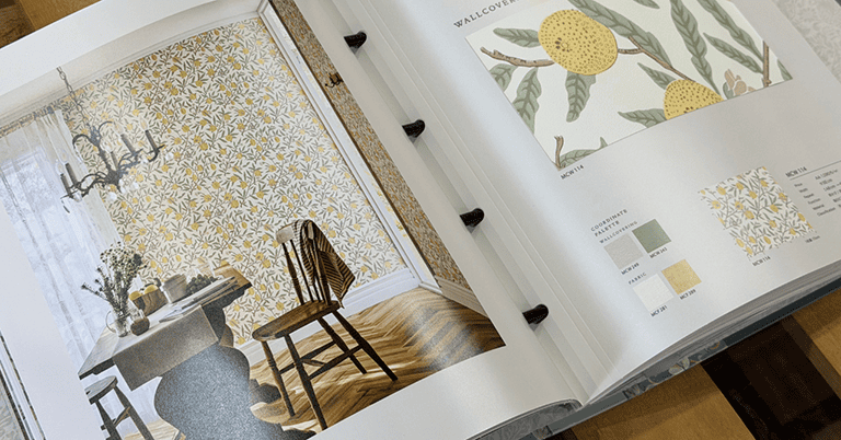 モリスの壁紙を使用したダイニングルームの施工イメージと、対応する壁紙サンプルを掲載したコーディネートブックの見開き。左ページには黄色い果実と葉柄の壁紙に囲まれた木製テーブルと椅子の室内写真、右ページには同柄の拡大サンプルとカラーバリエーション、品番情報が整理されている。