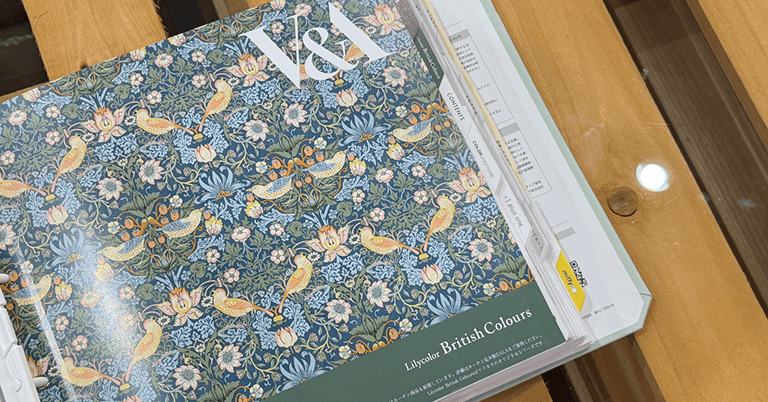 木製テーブルの上に置かれたモリス柄の壁紙サンプルブック。鳥と花が描かれたブルーグリーン系の『Lilycolor British Colours』デザインが表紙全面に広がり、上部には「V&A」のロゴ、右側には見出し付きのインデックスページが重なって見えている。