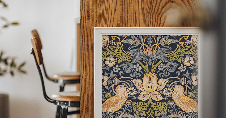 木目の柱に額装されて飾られたウィリアム・モリスの代表的な鳥と草花文様の壁紙デザイン。左右対称の構図で鳥と花が描かれ、背後にはぼかした椅子や室内空間が見え、インテリアアートとしての壁紙の魅力が伝わる。