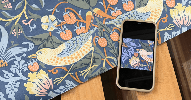 ウィリアム・モリスの『いちご泥棒』柄の壁紙サンプルの上にスマートフォンが置かれている様子。鳥や果実、草花が描かれたブルー系の文様が広がり、スマートフォン画面にも同じ壁紙柄が映り込み、デザインの細部とスケール感が伝わる構図。
