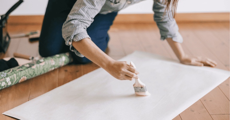 木目の床に広げた壁紙の裏面に刷毛でのりを塗っているDIY作業風景