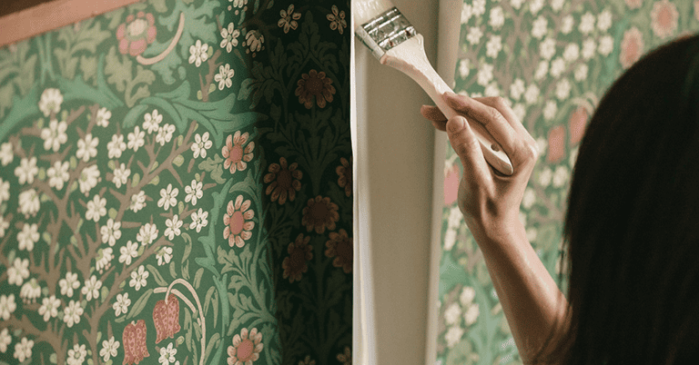 グリーン系のウィリアム・モリス調の草花といちご柄の壁紙をDIYで施工している様子。壁の角部分で、刷毛を使って壁紙の端や枠まわりに丁寧に糊を塗っており、手作業による貼り替え作業の雰囲気が伝わる。