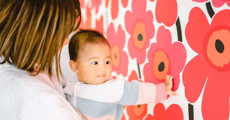 赤いマリメッコのウニッコ柄の壁紙に赤ちゃんが手を伸ばして触れている様子。抱っこした母親と一緒に大きな花柄デザインを見ている室内シーン