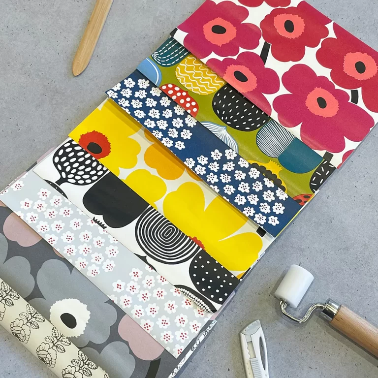 マリメッコのさまざまな柄の壁紙サンプルが重ねられ、ローラーとカッターが横に置かれている作業風景