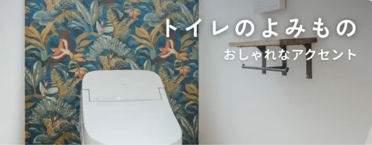 南国の植物柄の壁紙をアクセントにしたトイレ。白い便器と木製の棚があり、「トイレのよみもの おしゃれなアクセント」と文字が入っている。